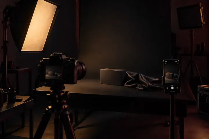 Estúdio de foto de produto: câmera no tripé, softbox acesa, caixa preta e tecido de cetim sobre fundo infinito, celular gravando ao lado.